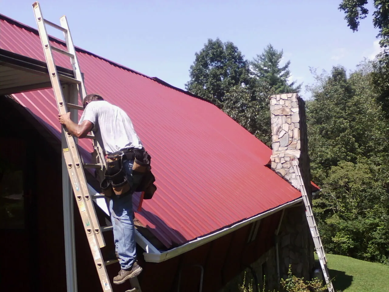 Roofer on a ladder beside a red metal roof and stone chimney — Soffit & Fascia Repair by Roofing Brothers in Bernalillo